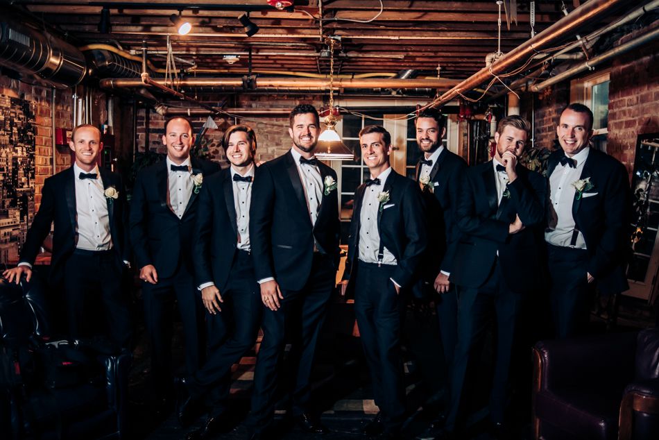 Groomsmen in black tuxedos pose together in an industrial-style venue with exposed pipes and brick walls.