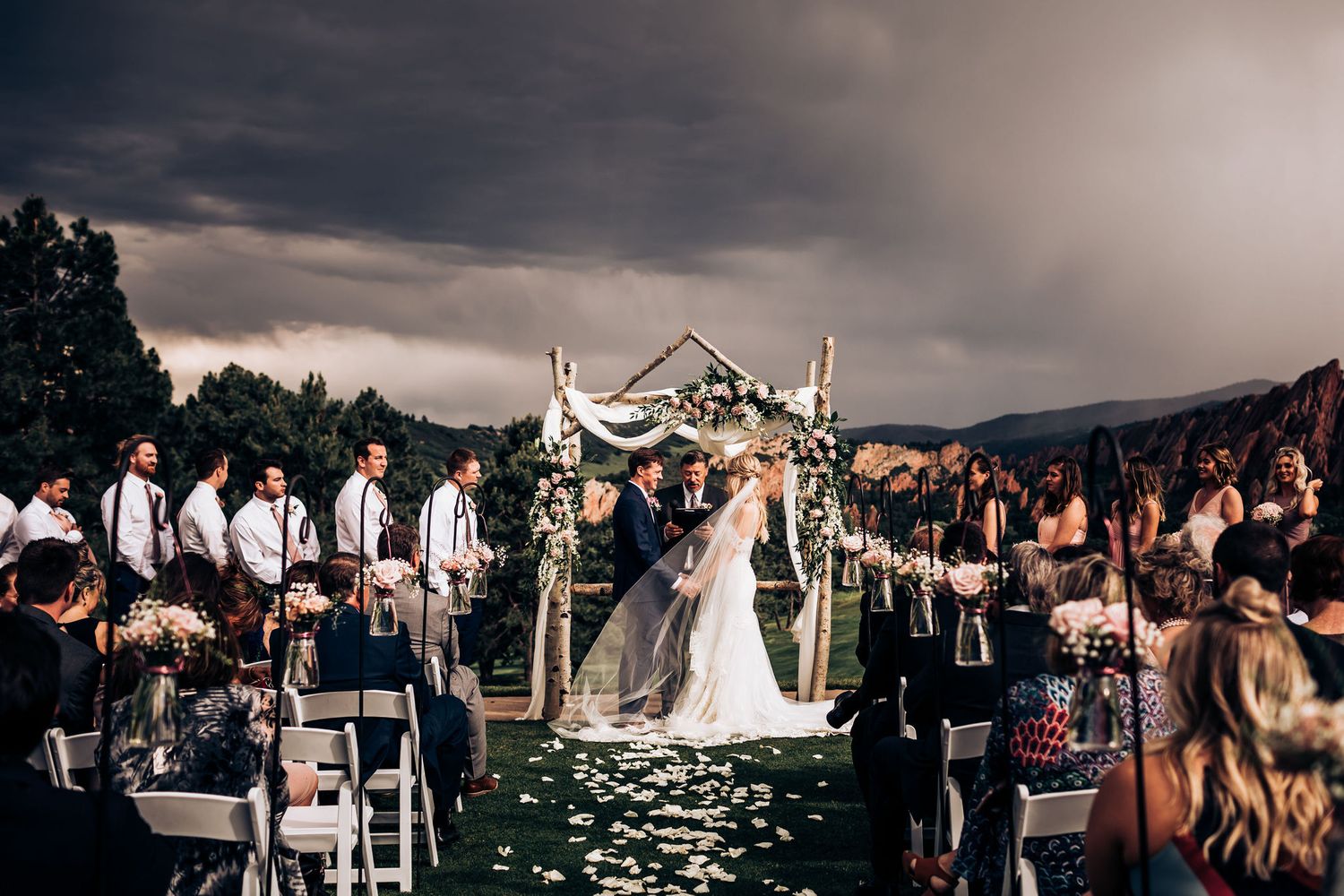 Dramatic evening wedding ceremony with guests seated outdoors under wooden arch and moody sky.