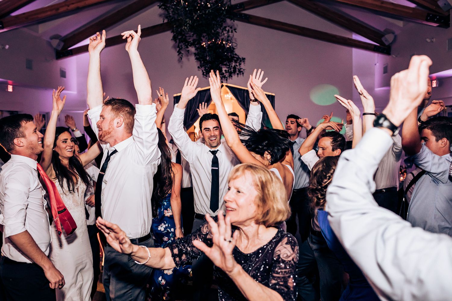 Crowd of wedding guests dance with hands raised during reception celebration.