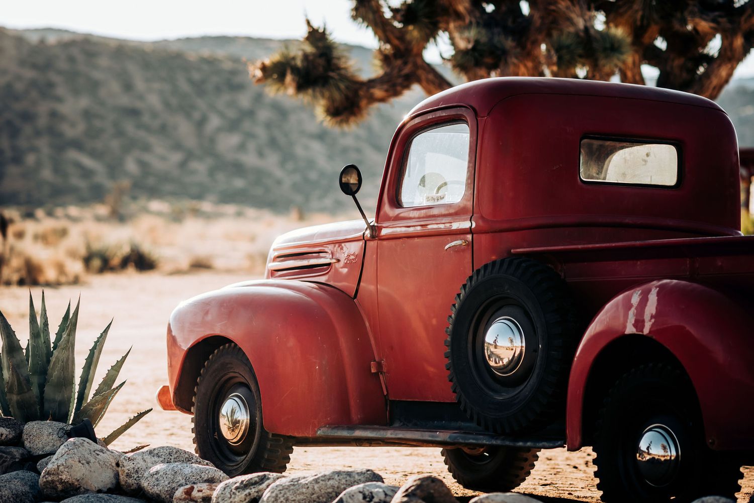 Vintage red pickup truck parked in desert landscape with Joshua trees and mountains.