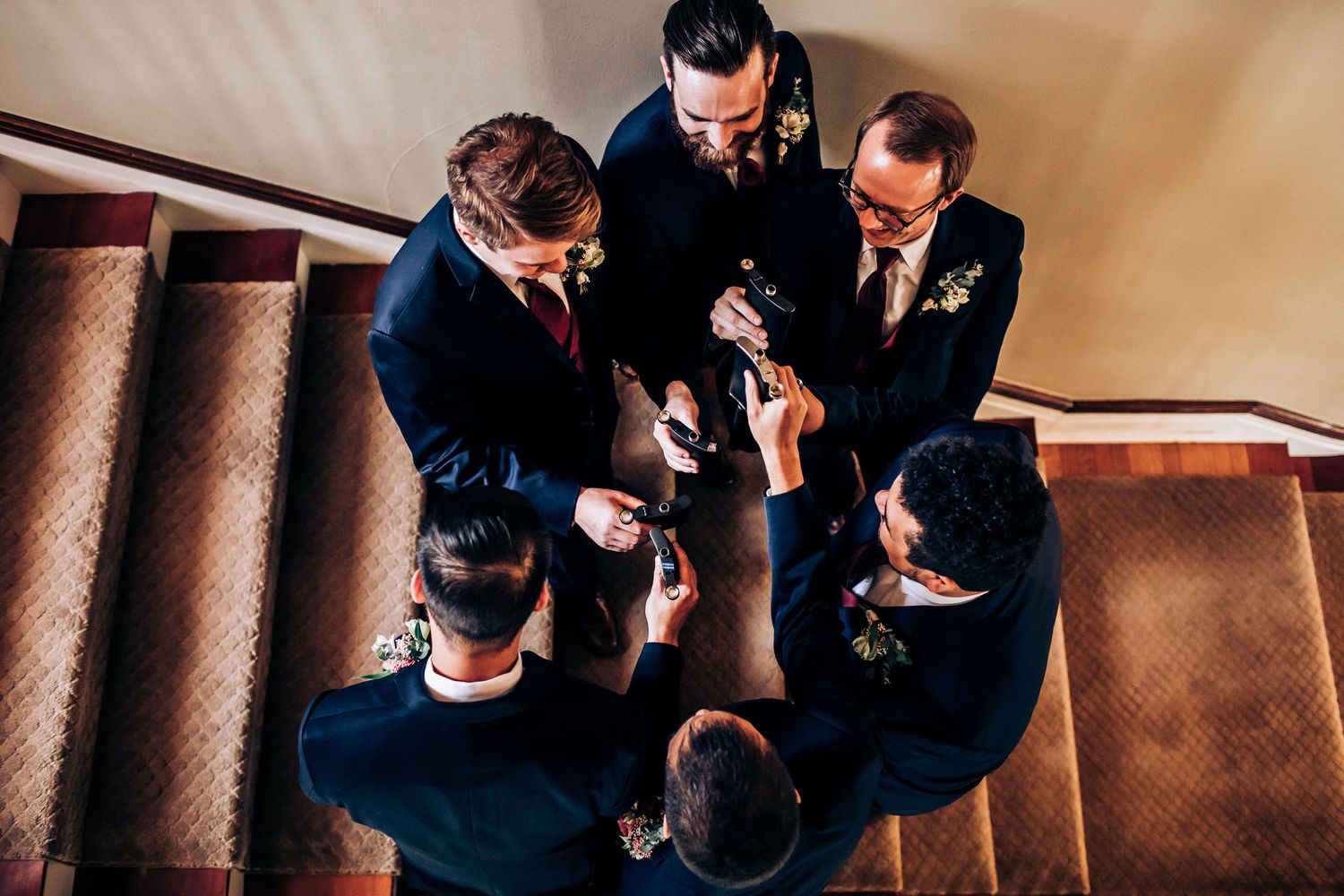 Groomsmen gathered in a circle on carpeted stairs having drinks before the wedding ceremony.