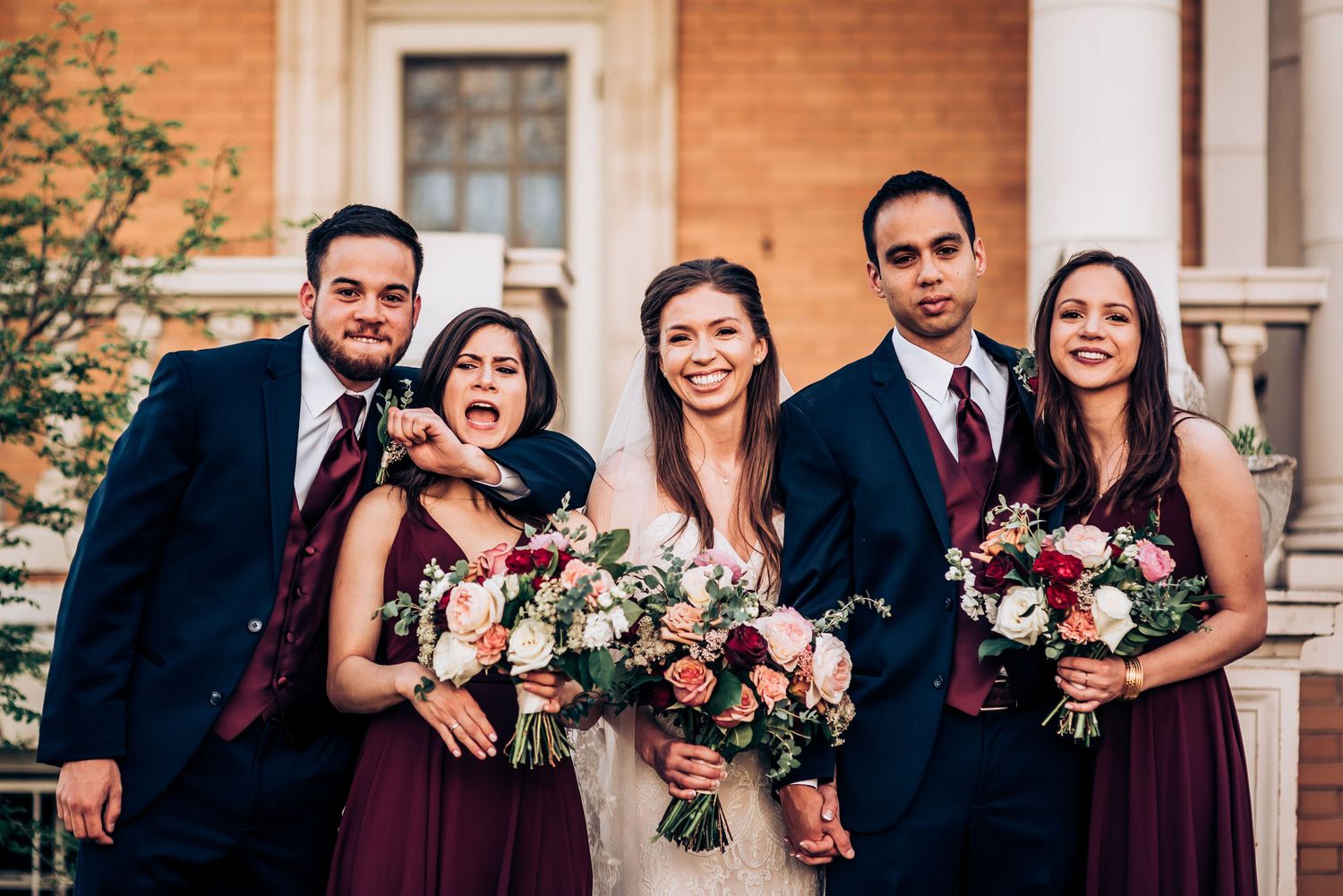 Wedding party with burgundy dresses holding floral bouquets poses together outside brick building.