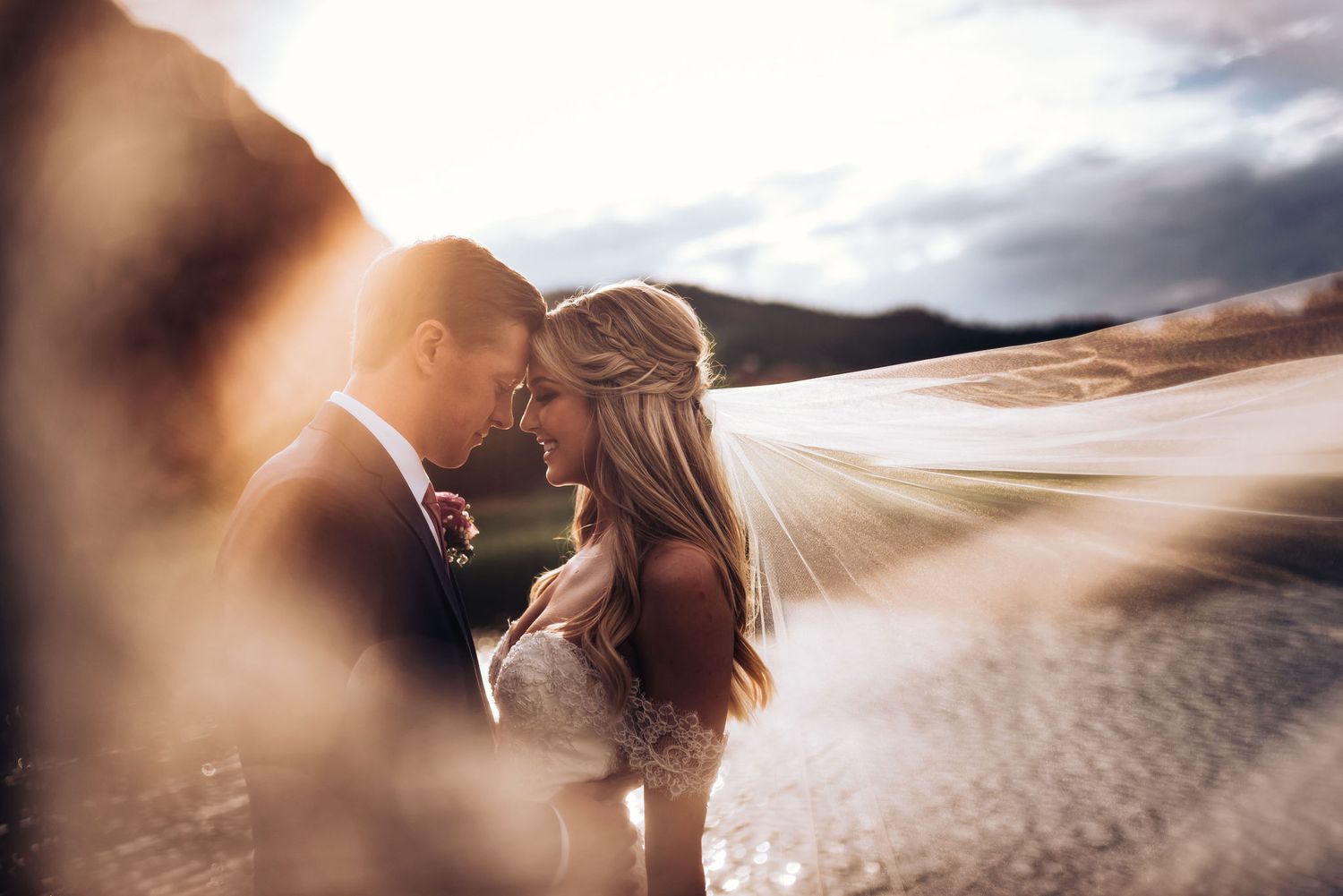 Artistic wedding portrait showing a couple embracing with flowing veil and dramatic clouds in background.