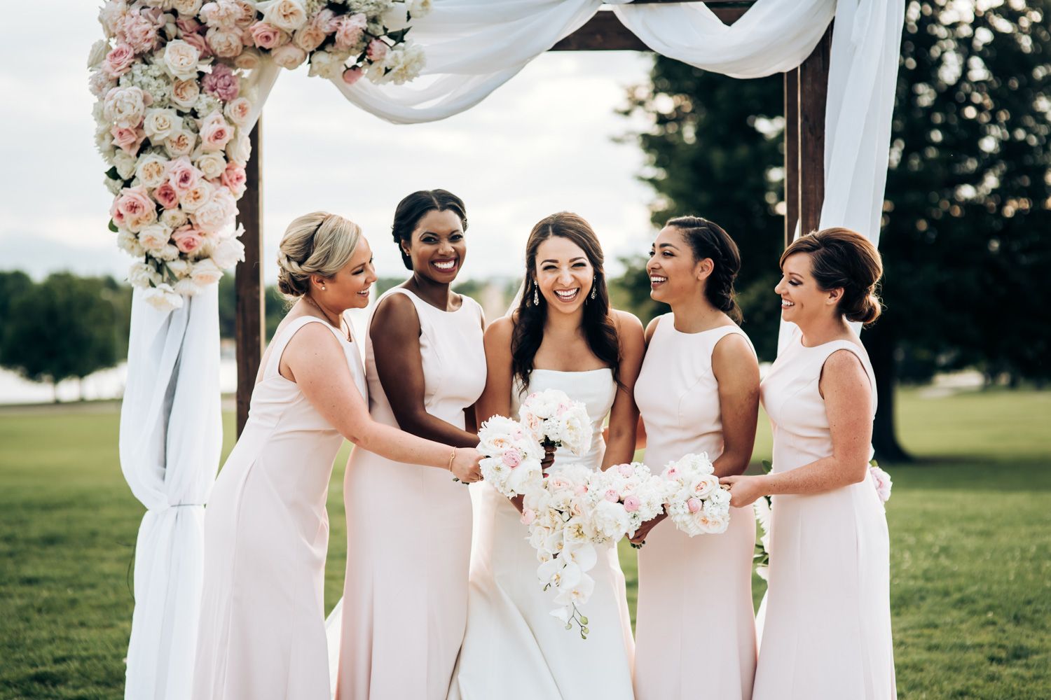 Bridesmaids in blush pink dresses celebrate with bride under floral arch outdoors.