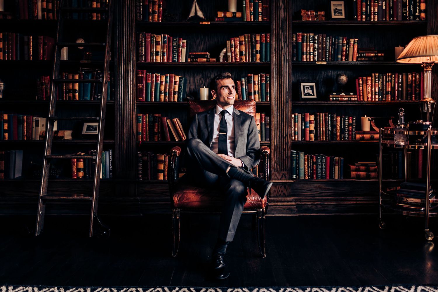 Person sitting in elegant library setting with dark wooden bookshelves and vintage lamp.