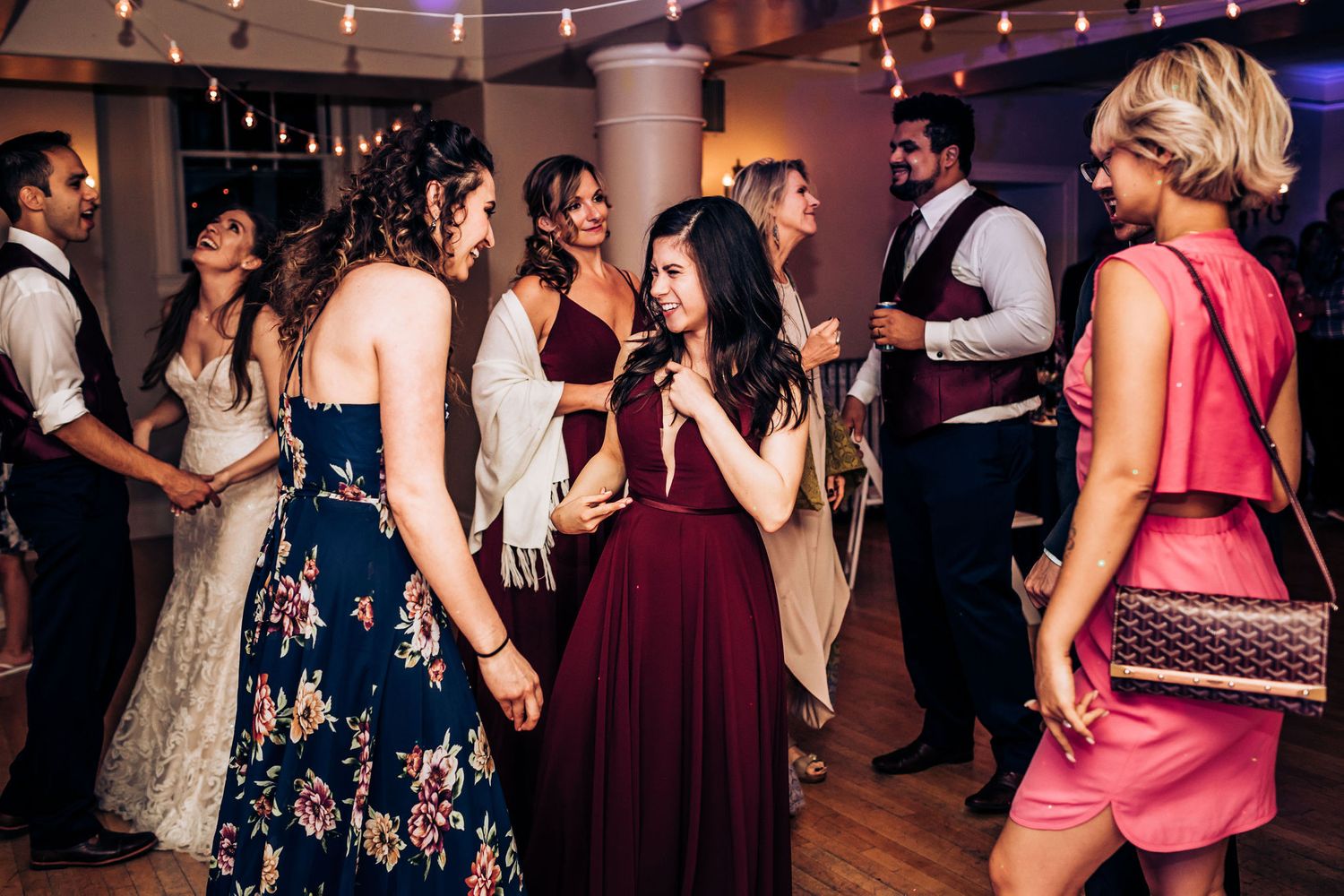 Party guests in formal attire socialize and dance at an evening celebration with string lights overhead.