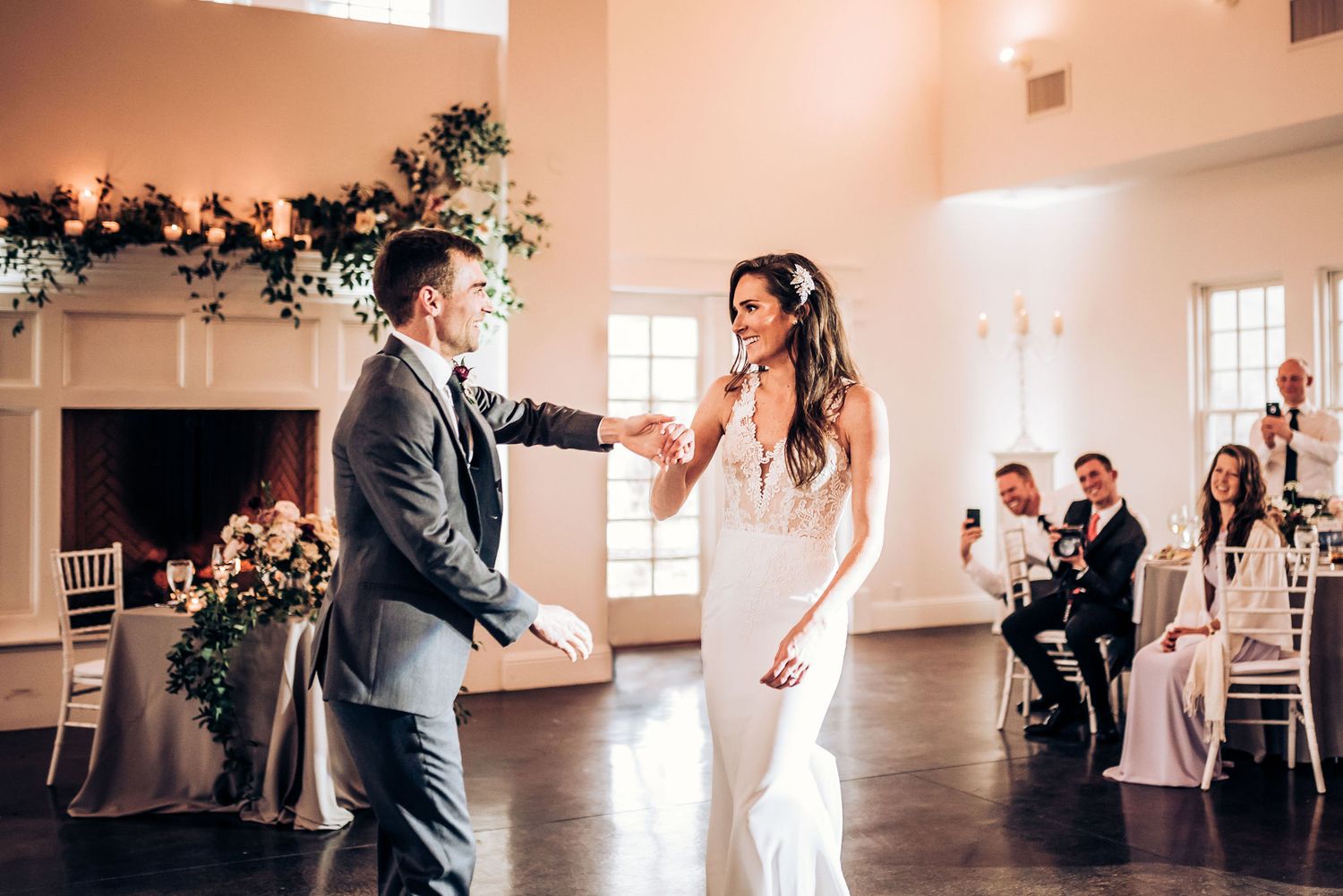 A couple shares their first dance in an elegant white room with greenery decorations and watching guests.