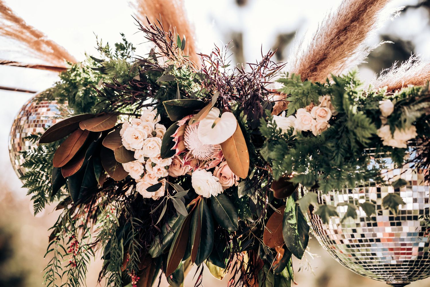 Elegant floral arrangement with magnolia leaves, pampas grass, and blush flowers hanging from a wedding arch.