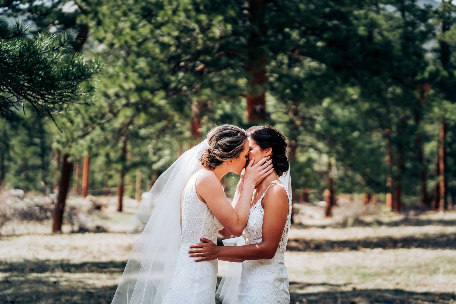 An intimate embrace between two brides in white wedding dresses in a forest setting.