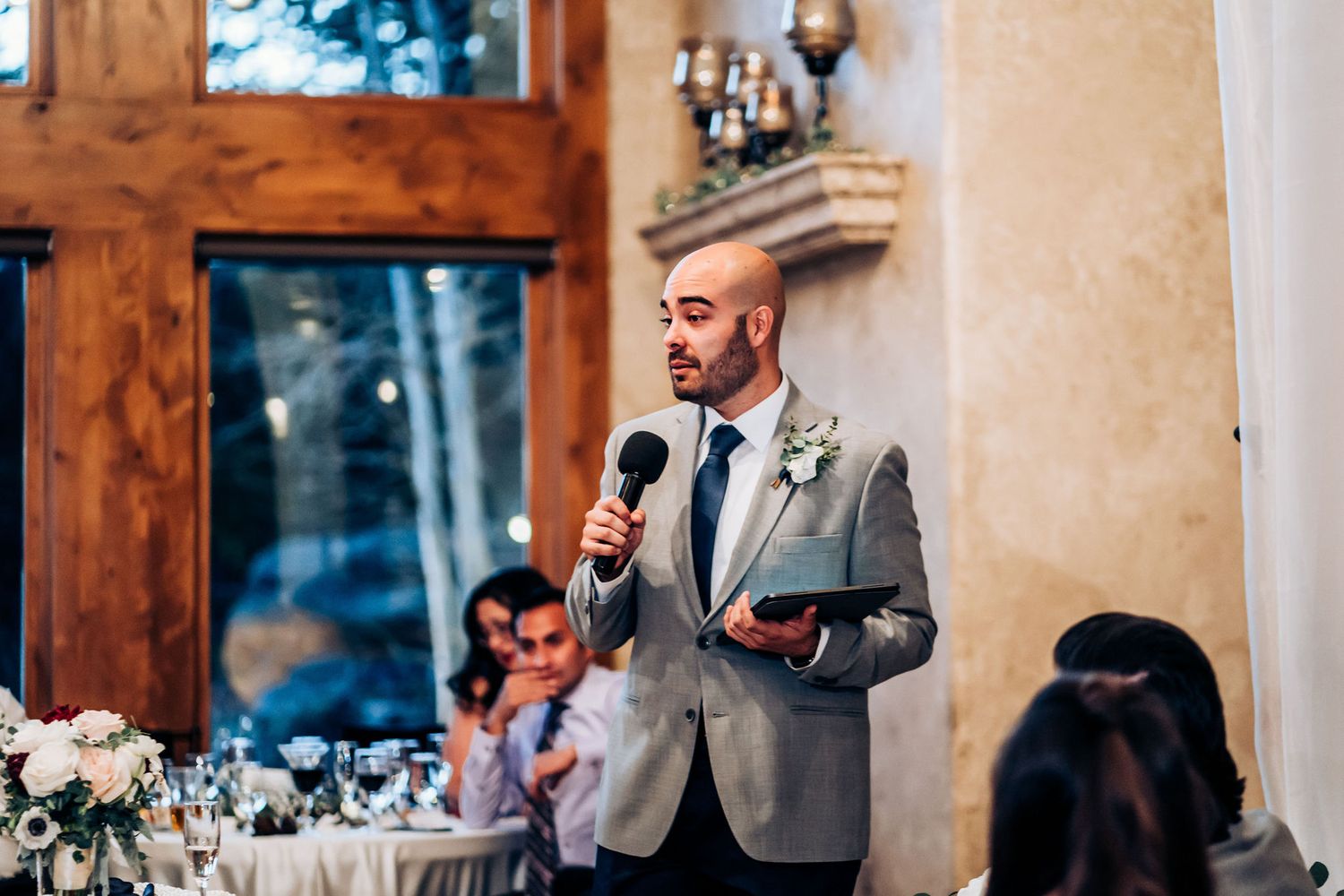 Person in grey suit delivering wedding speech while holding microphone at reception.