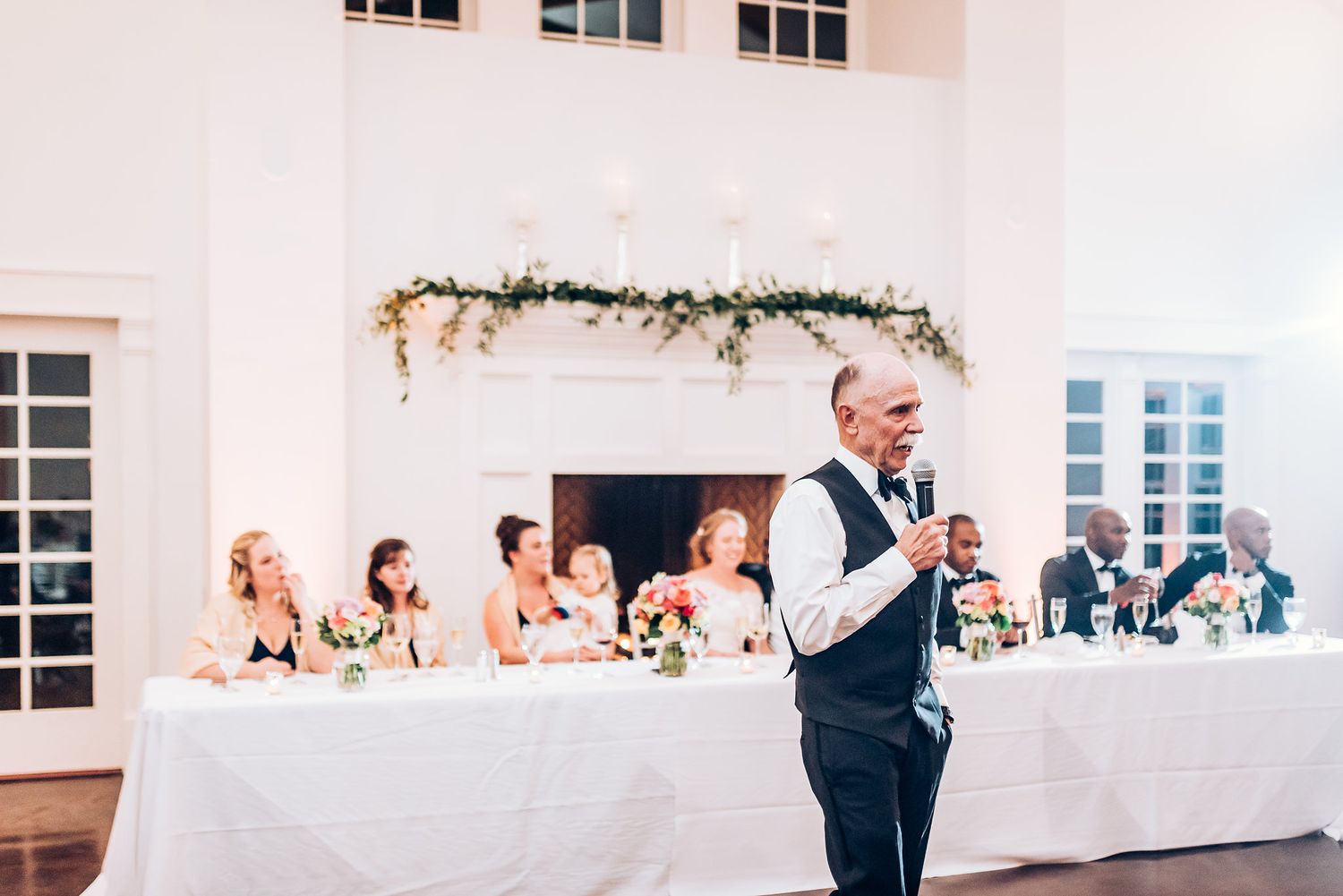 Speaker delivers wedding toast in front of decorated head table at elegant venue.