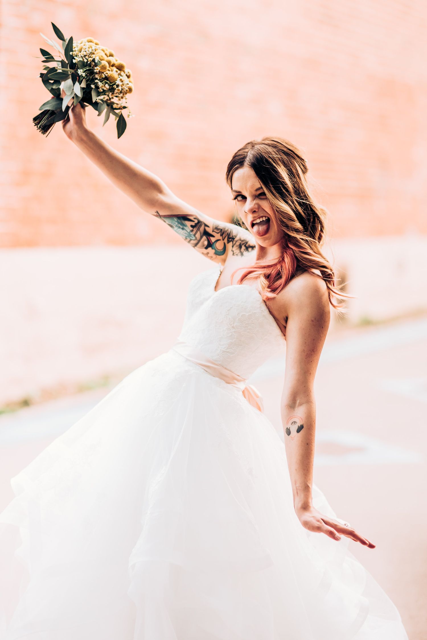Joyful bride in white ballgown dress holding bouquet against peach wall background.