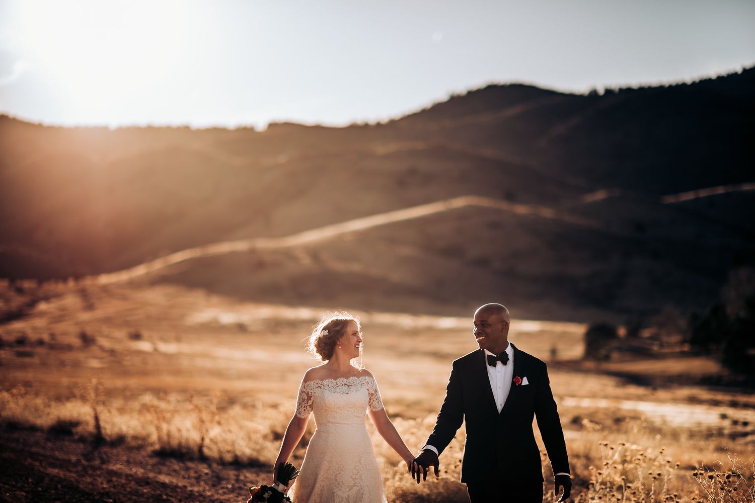 Couple holds hands while walking along hilltop during sunset with mountain views.