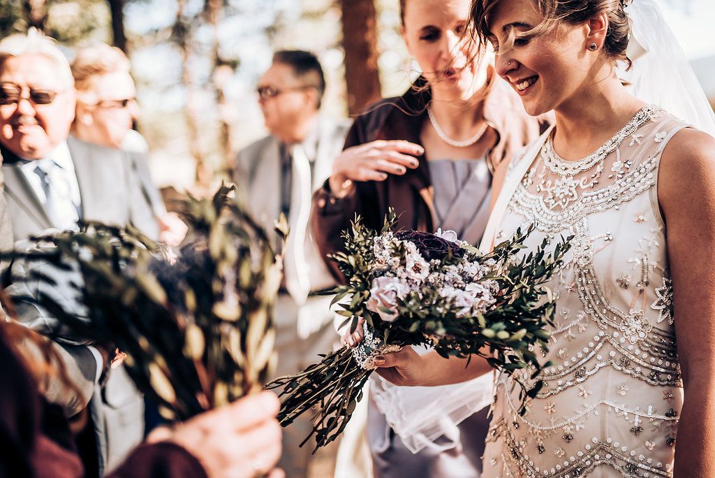 Wedding party admires a rustic bouquet with purple flowers during an outdoor forest celebration.
