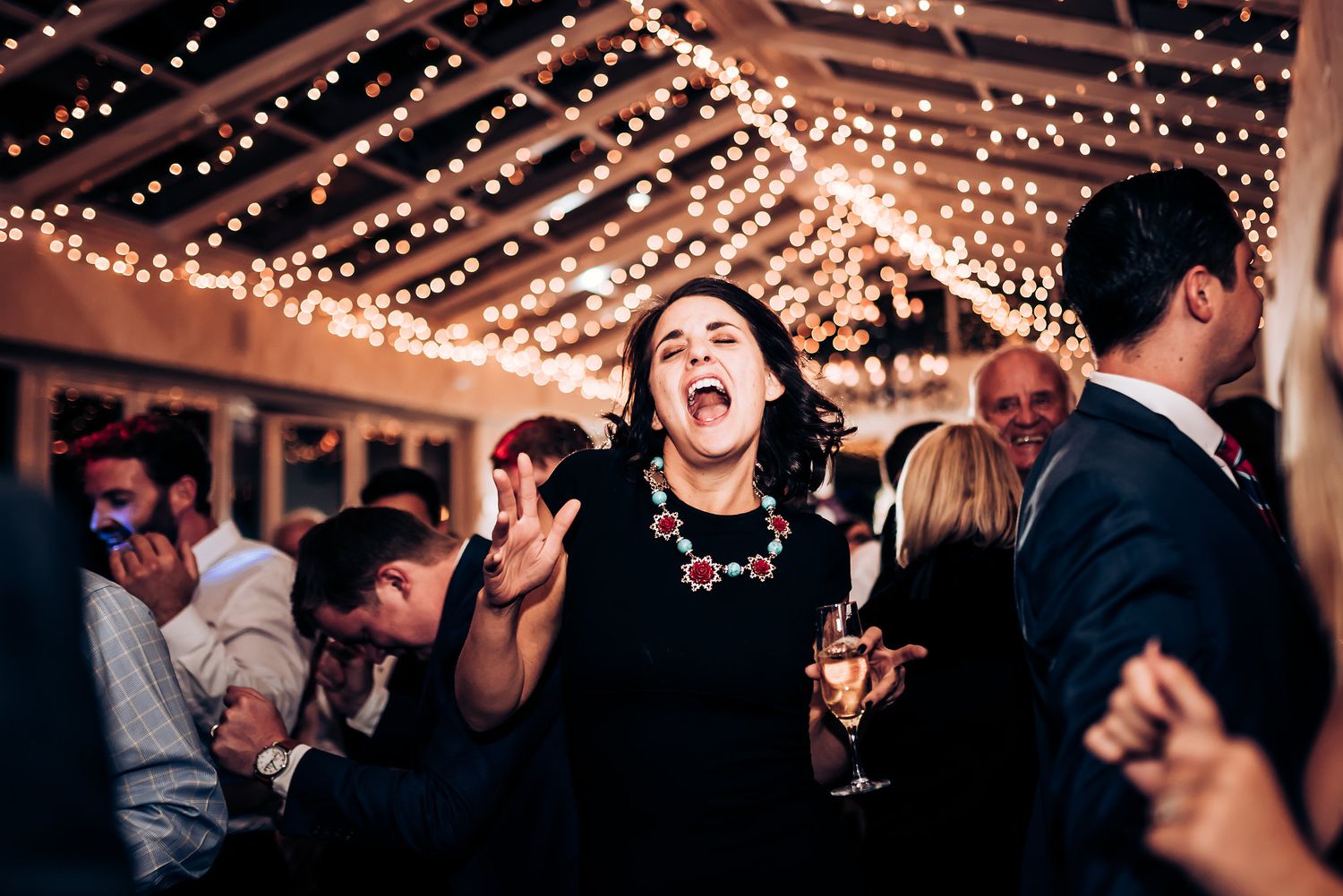 A guest in black dress sings enthusiastically under string lights at a celebration.