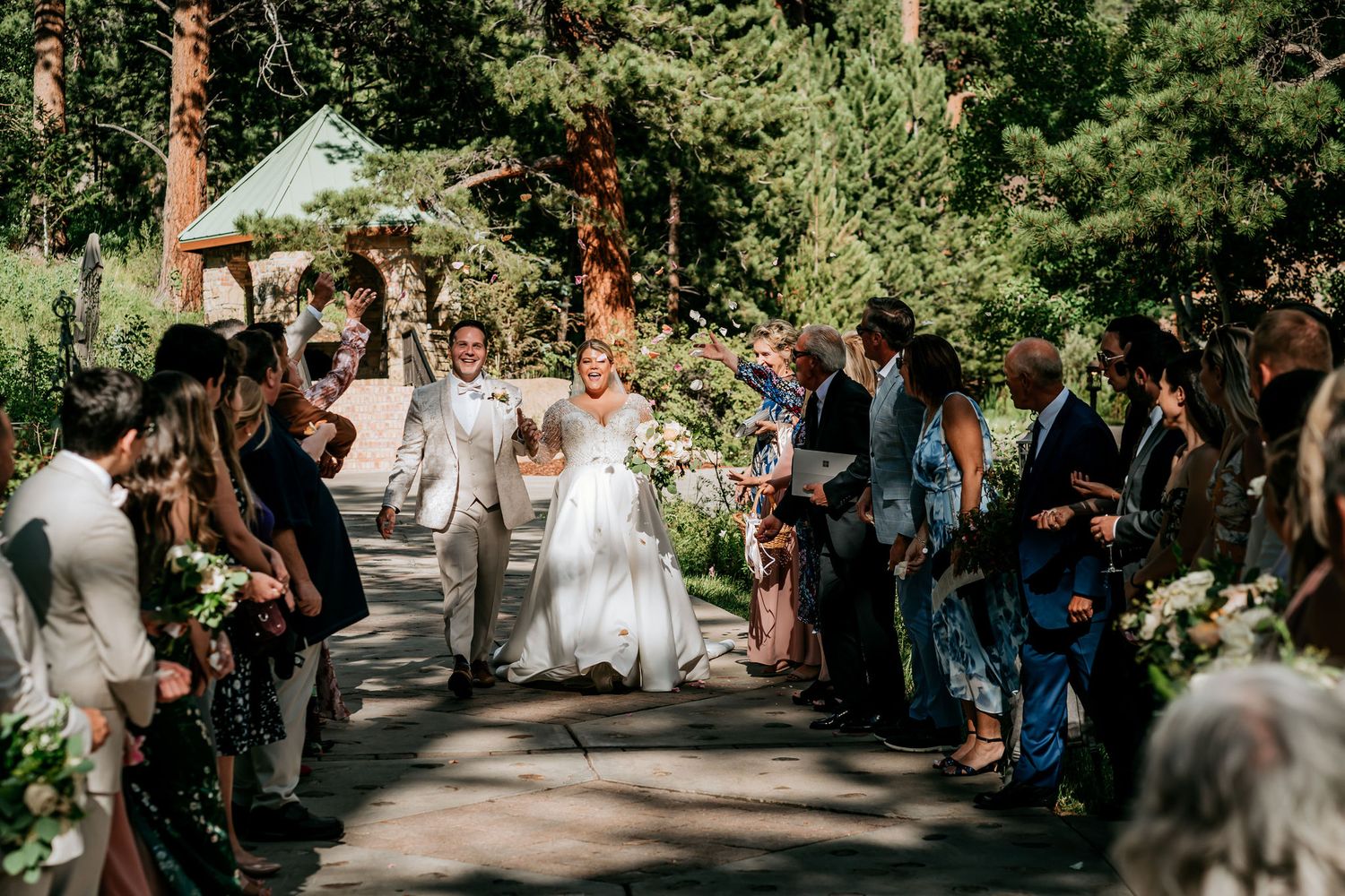 Wedding procession on a tree-lined path with guests watching from both sides.