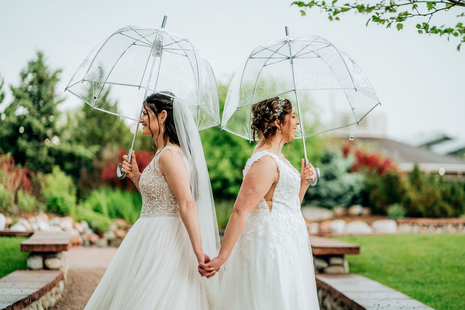 Two brides holding clear umbrellas in a garden setting.
