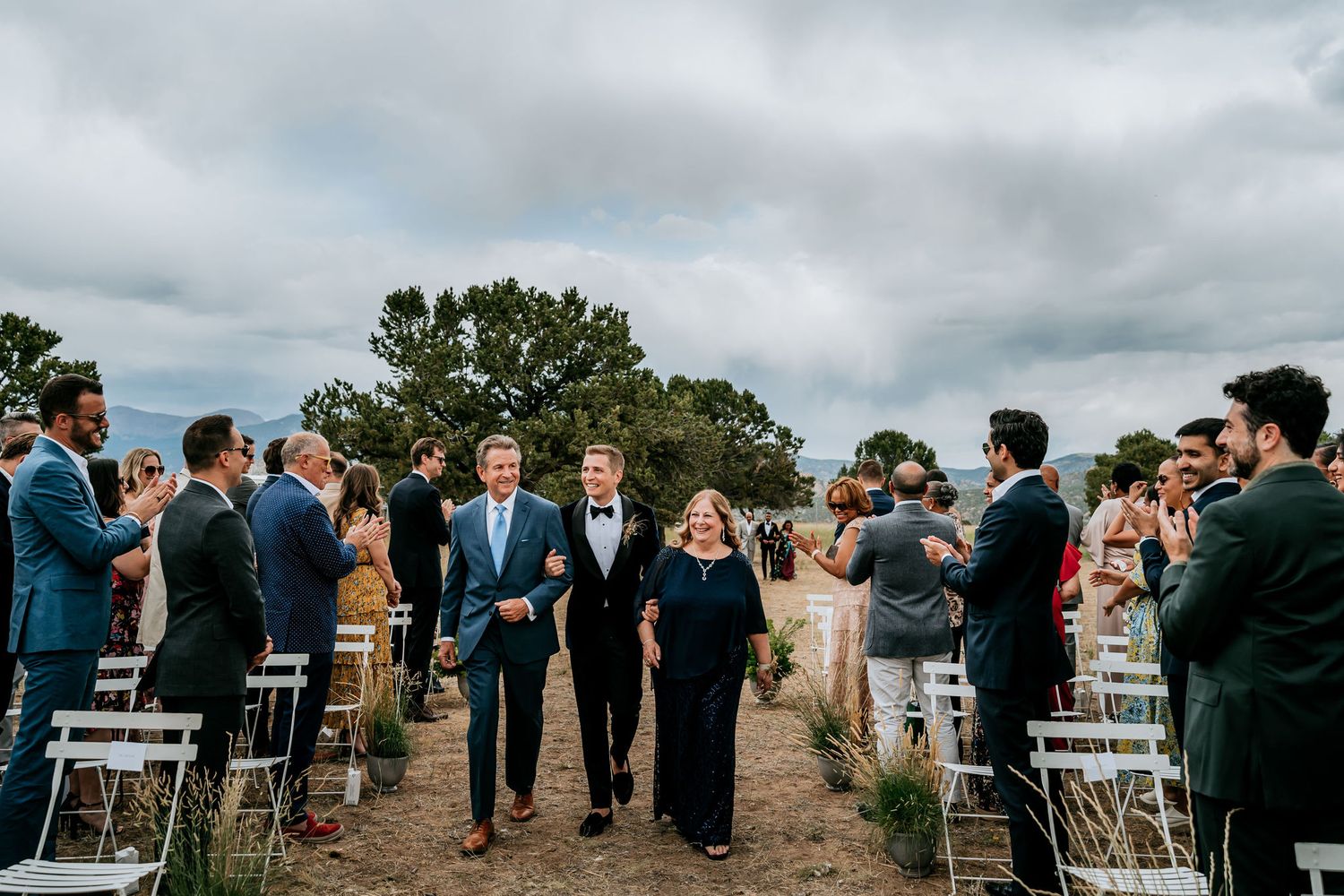 Wedding guests line an outdoor aisle as attendants walk through during a scenic mountain ceremony.
