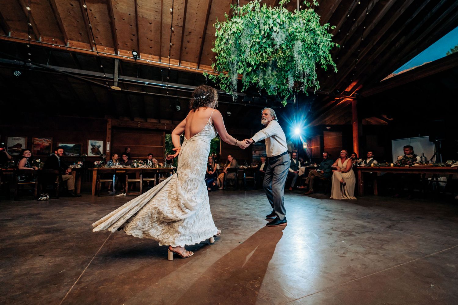 A wedding couple dances in a wooden barn venue lit by overhead string lights.