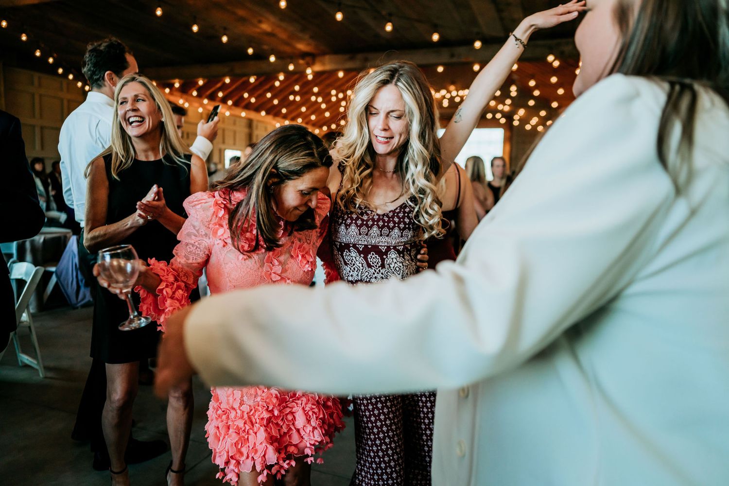 People dancing and celebrating at reception under warm string lights in rustic venue.