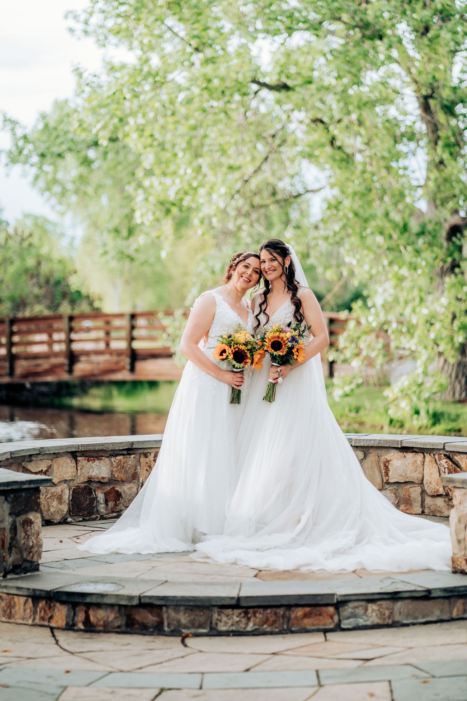Two brides holding sunflower bouquets pose on a stone bridge in a garden.