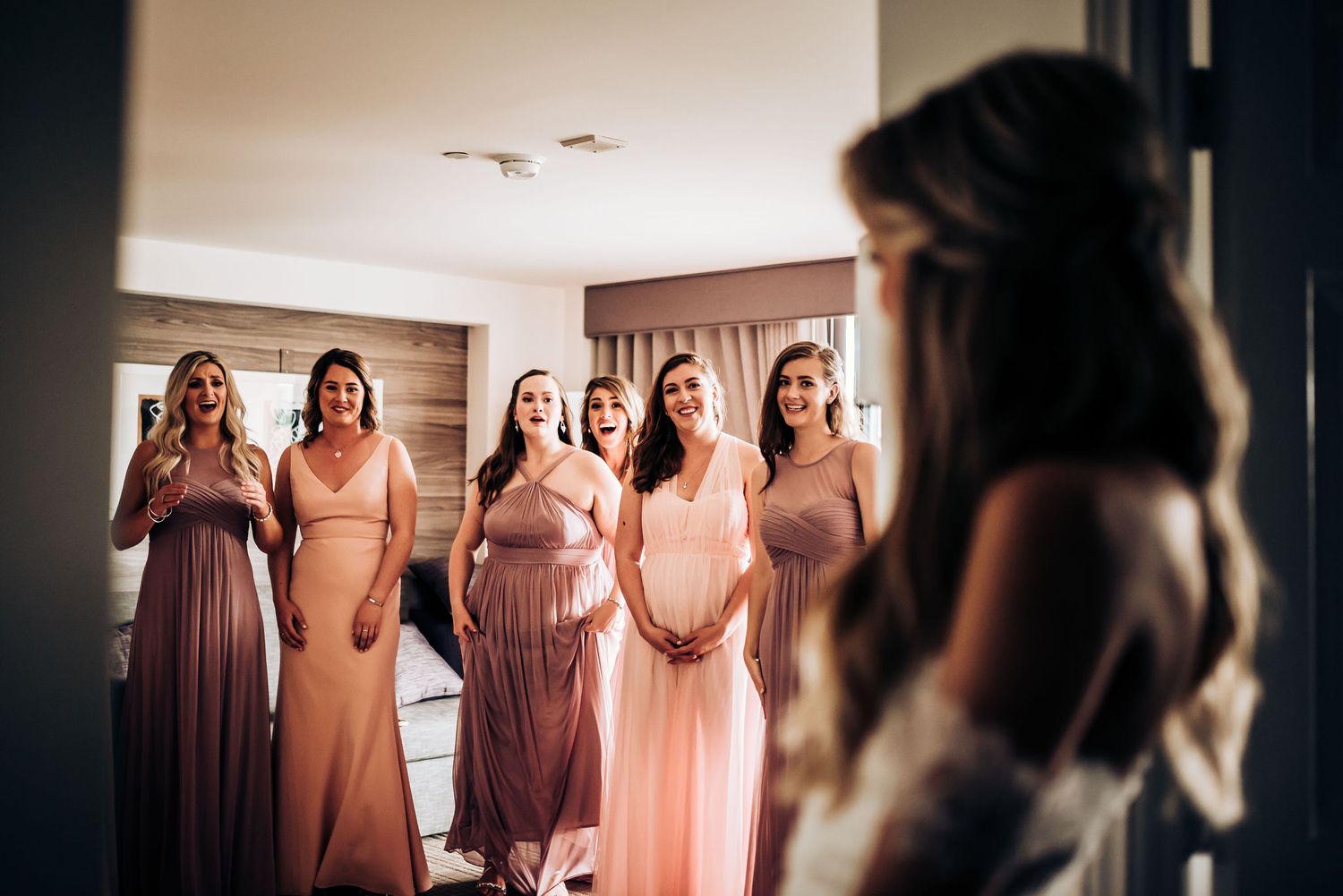 Bridesmaids in flowing pink dresses react with joy during a wedding reveal moment.
