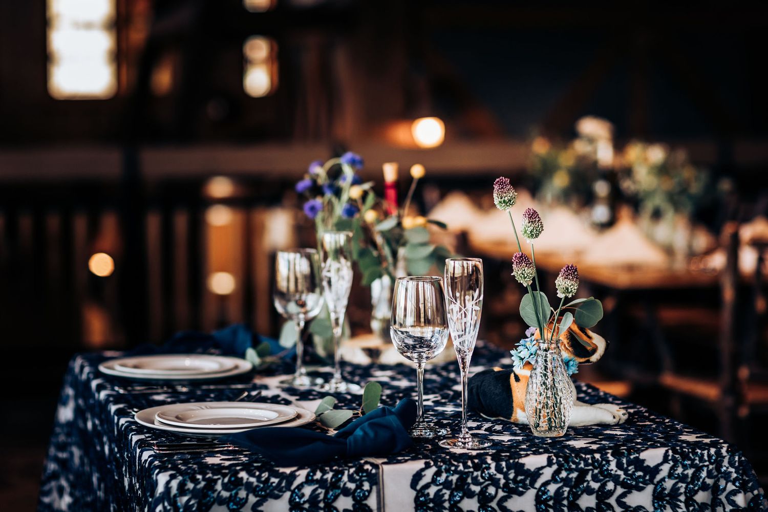 Elegant wedding reception table setting with wine glasses and floral centerpieces on navy patterned tablecloth.