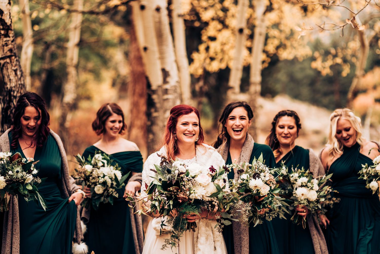 Wedding party in emerald green dresses laughs together while holding white floral bouquets in autumn setting.