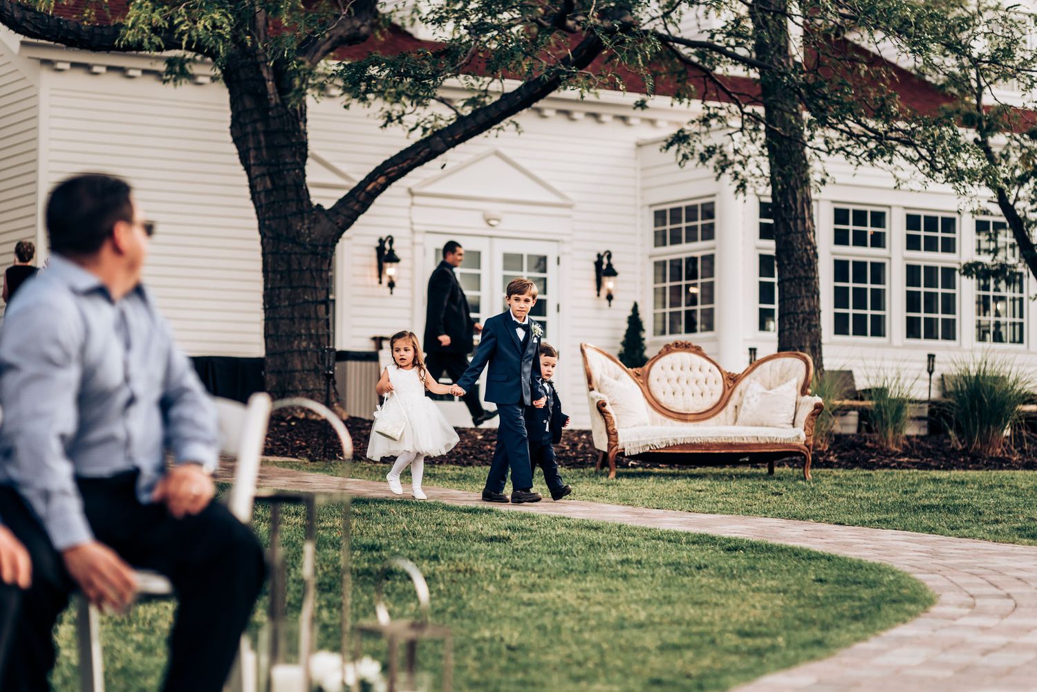 Wedding ceremony setup on a lawn outside a white building with guests gathering around decorative furniture.