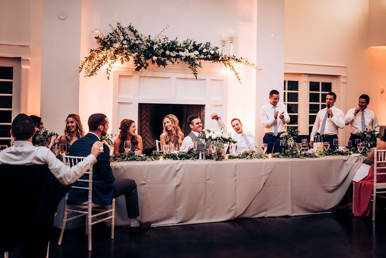 Reception venue features elegant head table with floral arrangements and white linens under soft lighting.