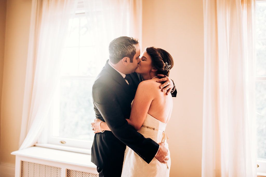 Romantic indoor wedding photography with soft window lighting and an intimate embrace.