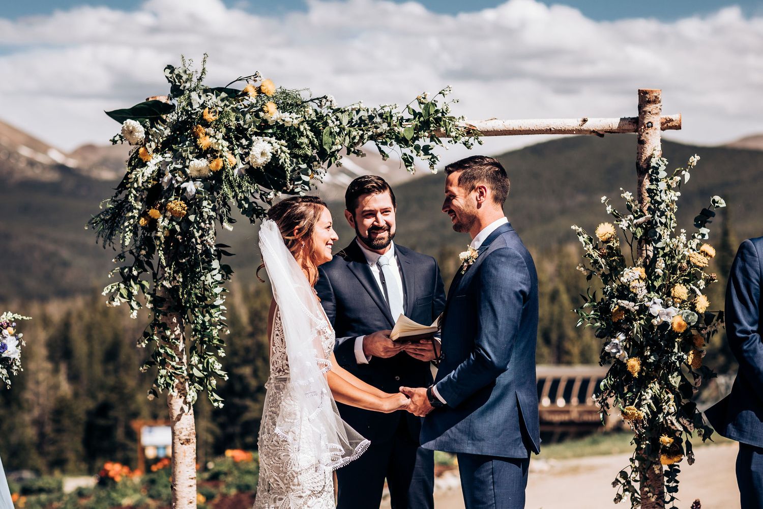 Wedding ceremony under floral arch with mountains and evergreen trees in background.