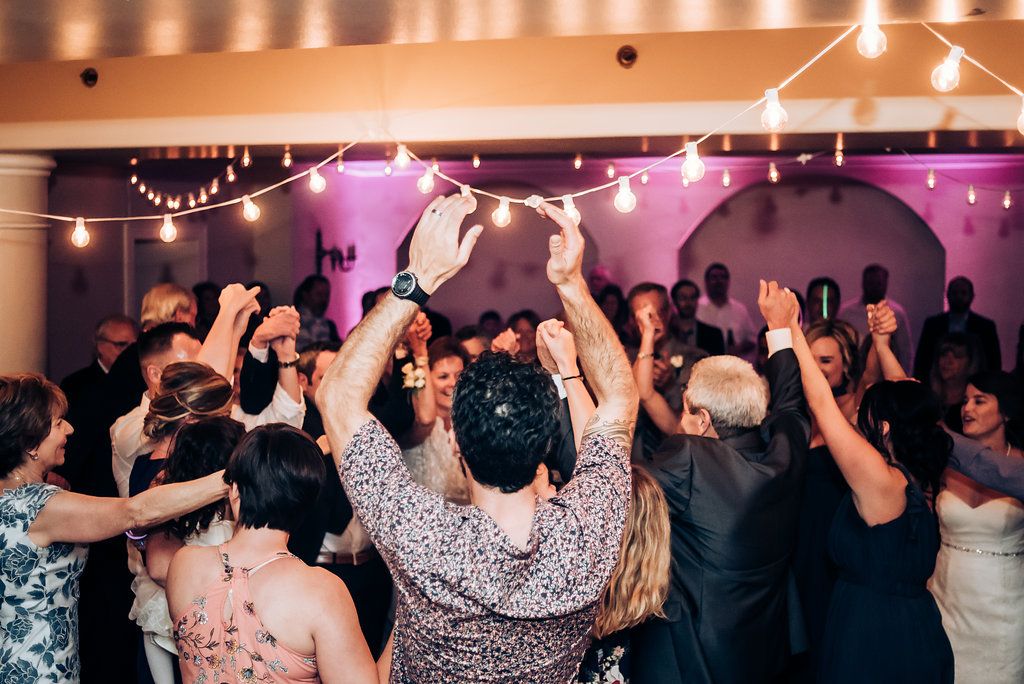 Dancing wedding guests raise their hands in celebration under string lights and purple decor.