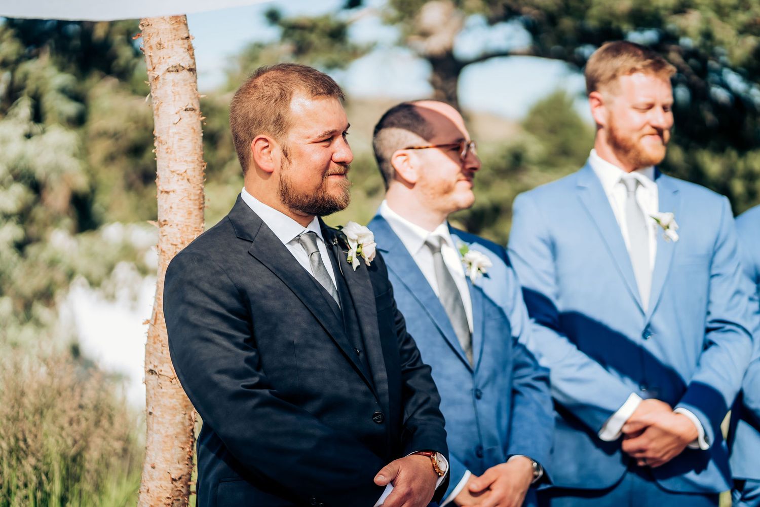 Three groomsmen in suits stand together during outdoor ceremony.