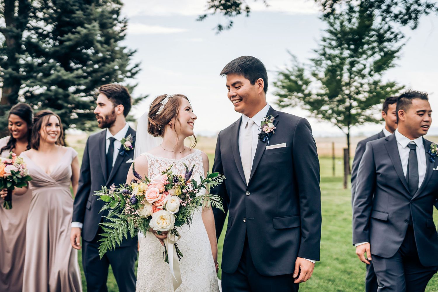 Wedding party enjoying a candid moment outdoors in a garden setting with trees and mountains in background.