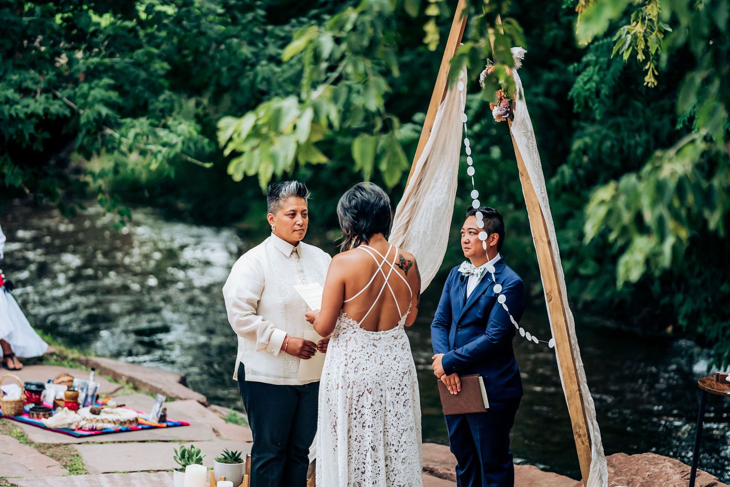 Outdoor wedding ceremony by a stream with wooden triangle arch and white decorations.