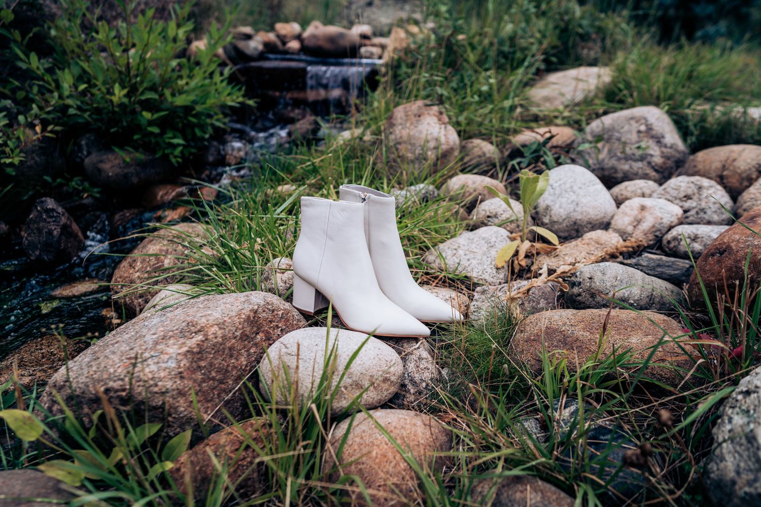 White leather ankle boots sit on rocky terrain near a small stream.
