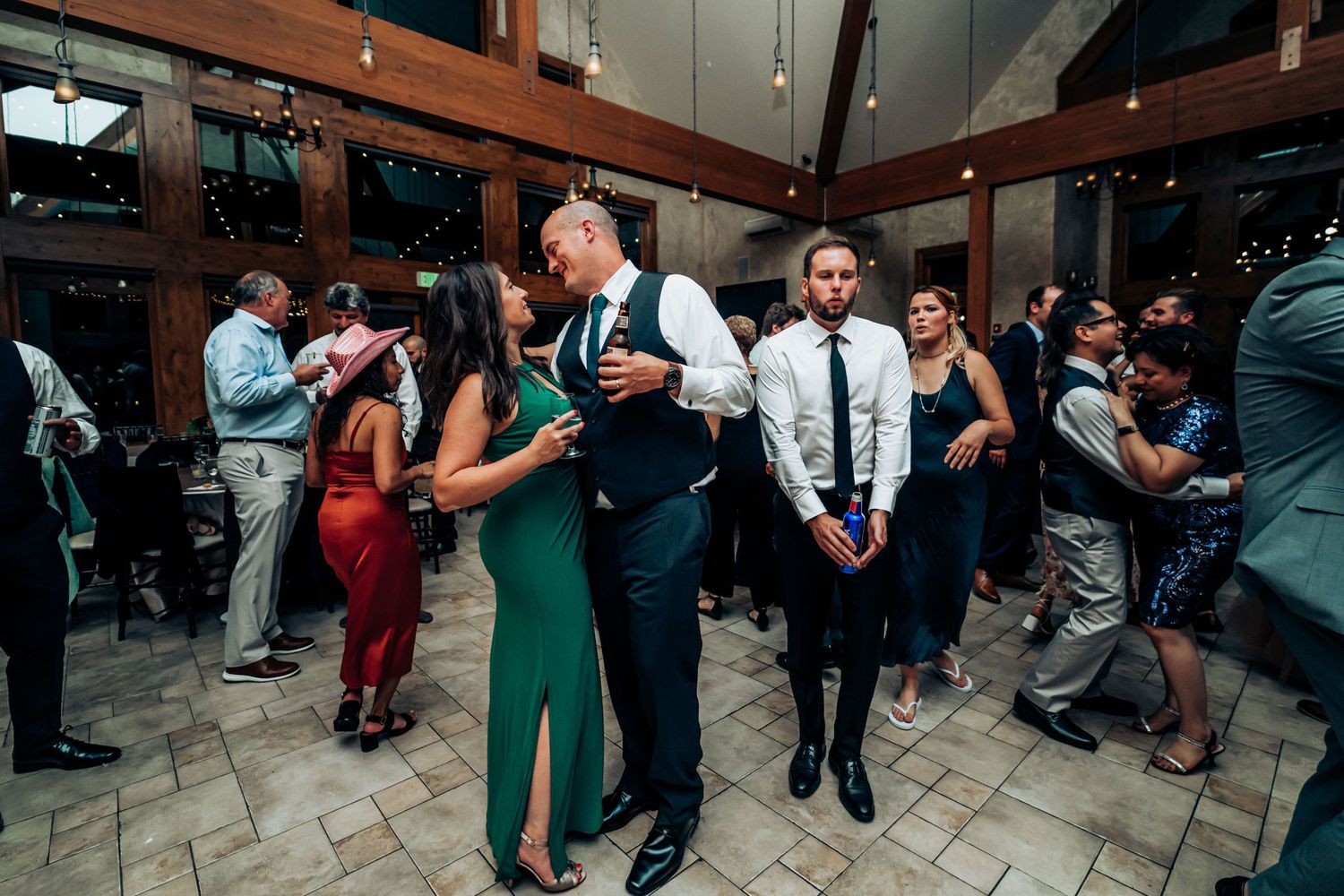 Party guests dancing and mingling at wedding reception in rustic venue with exposed wooden beams.