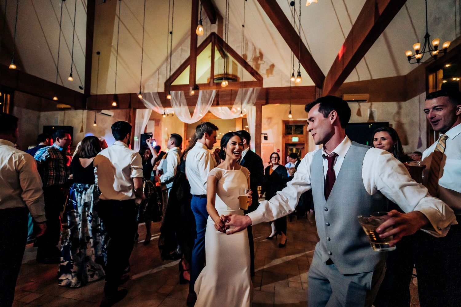 Party-goers celebrate on a dance floor in a rustic reception hall with wooden beams overhead.