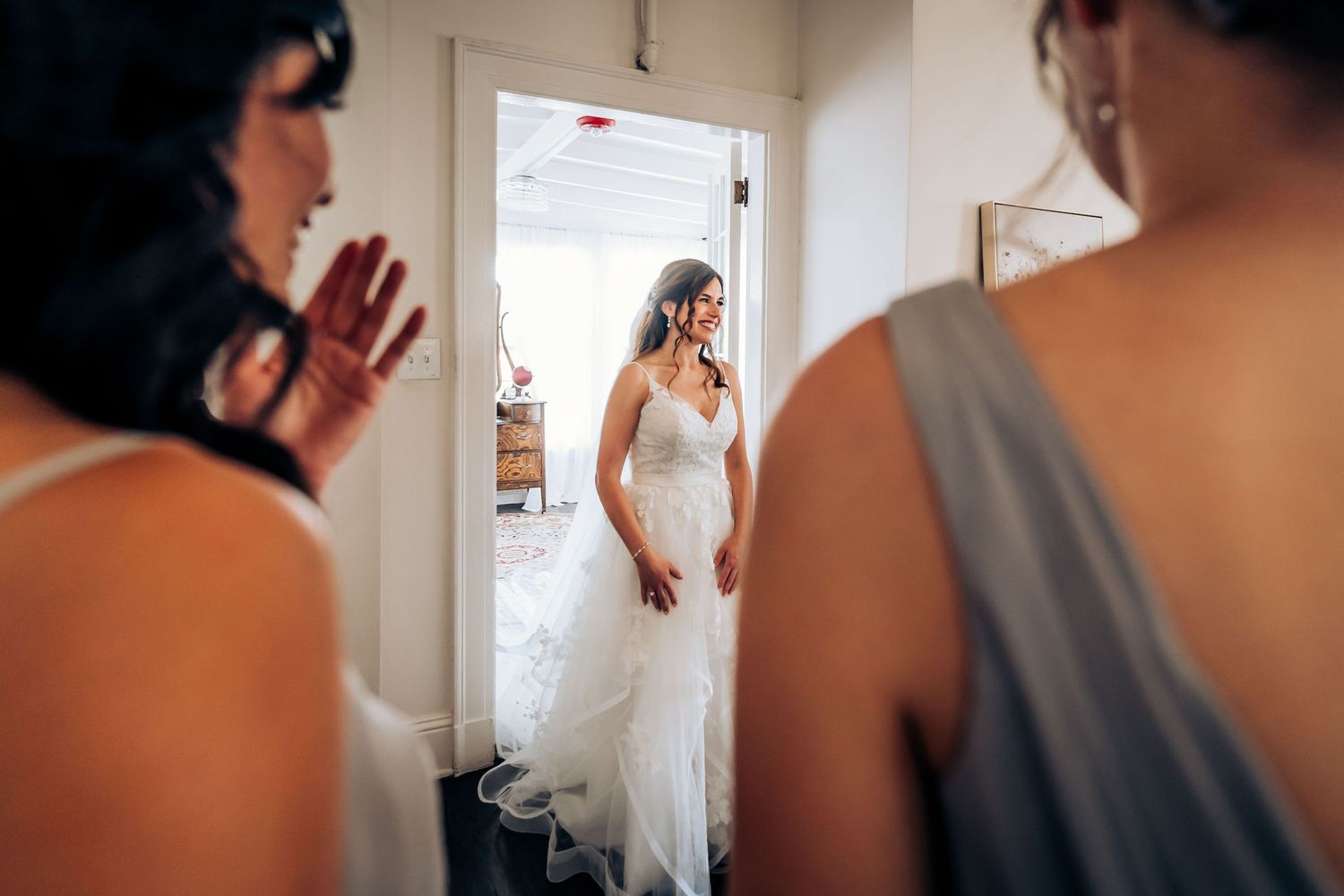A bride in a white wedding gown is revealed to her bridal party in a doorway.