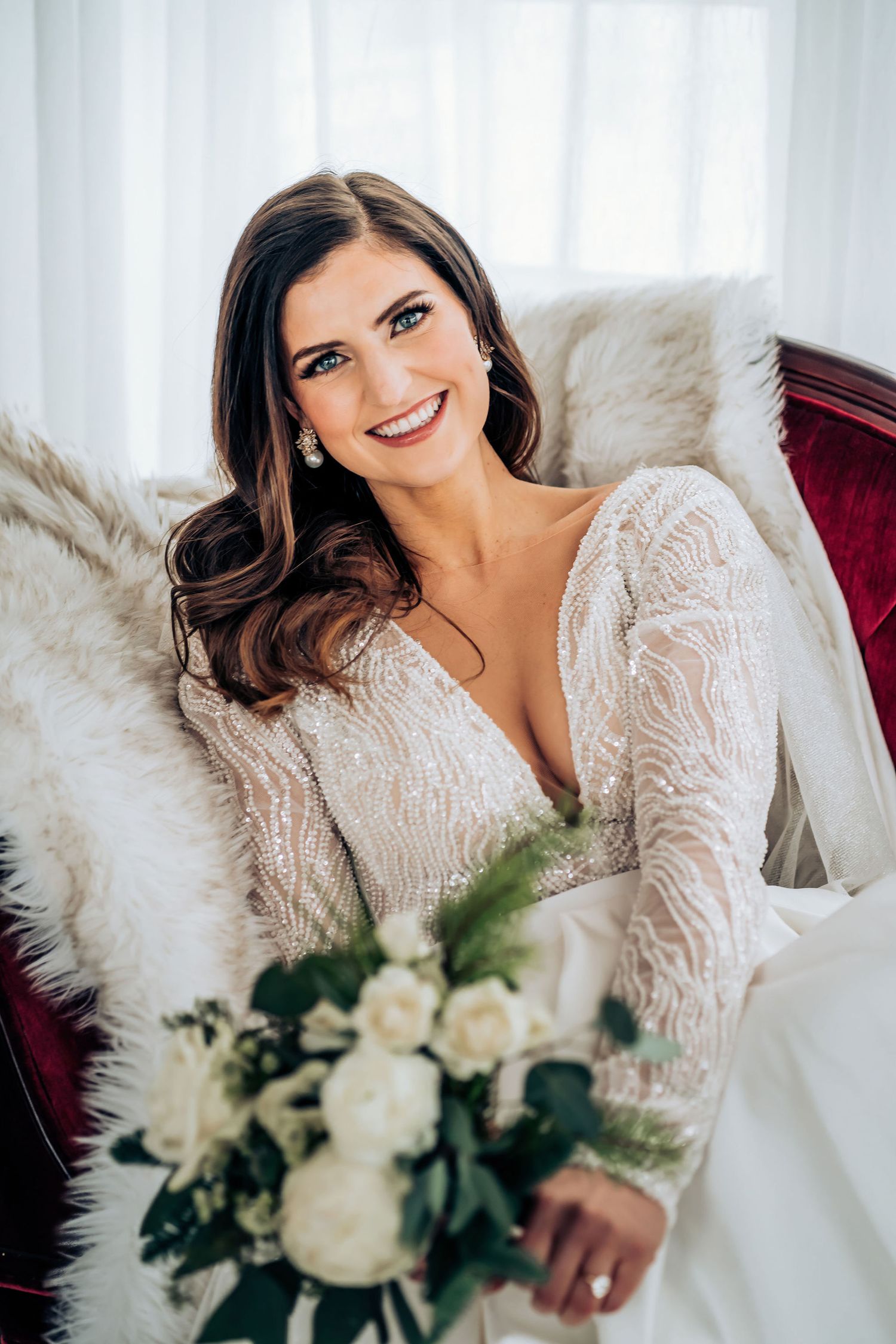 A bride in a lace long-sleeve wedding dress holds a white bouquet while sitting on a velvet couch.