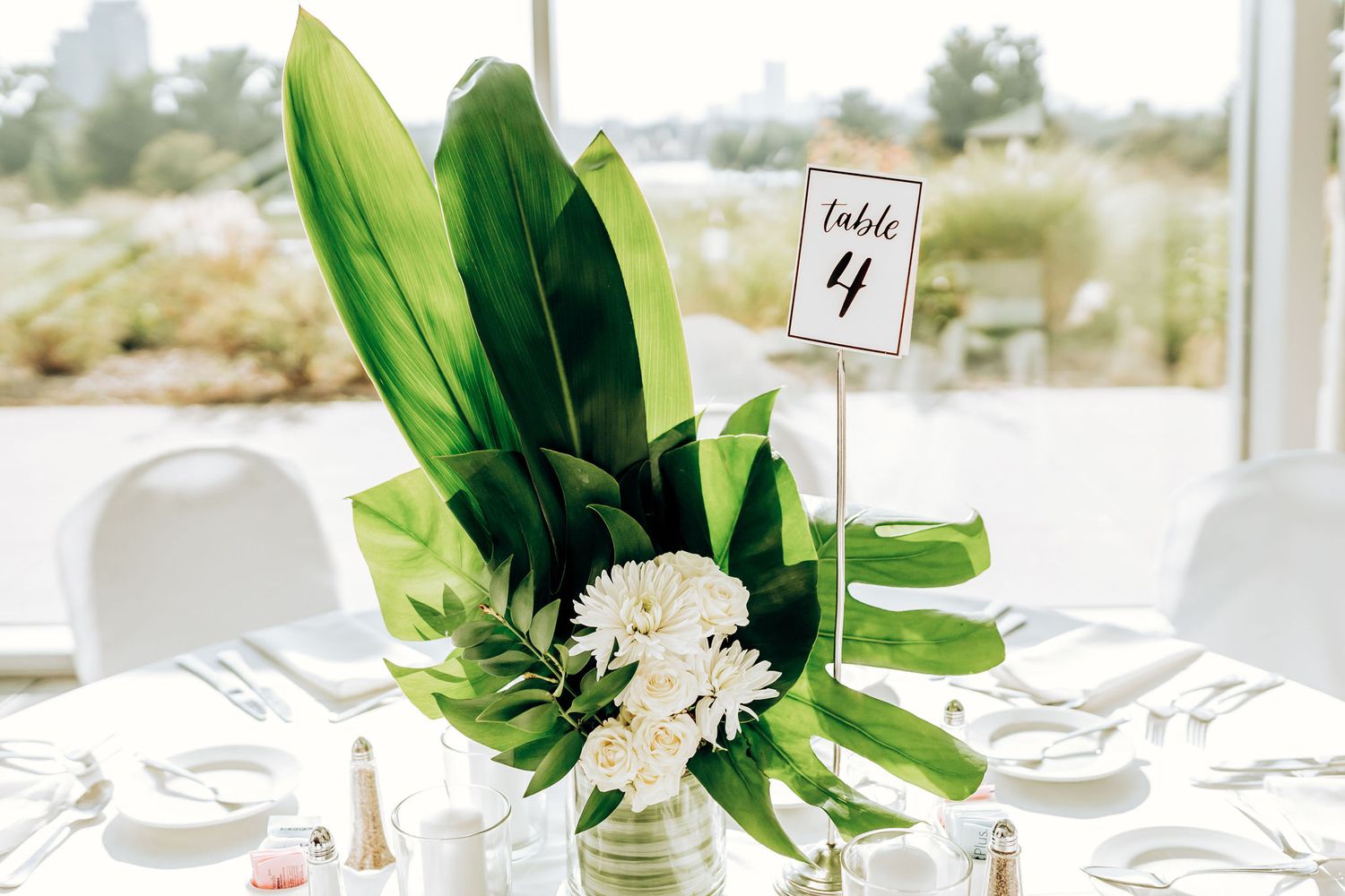Modern tropical wedding centerpiece with green leaves and white flowers on table number 4.