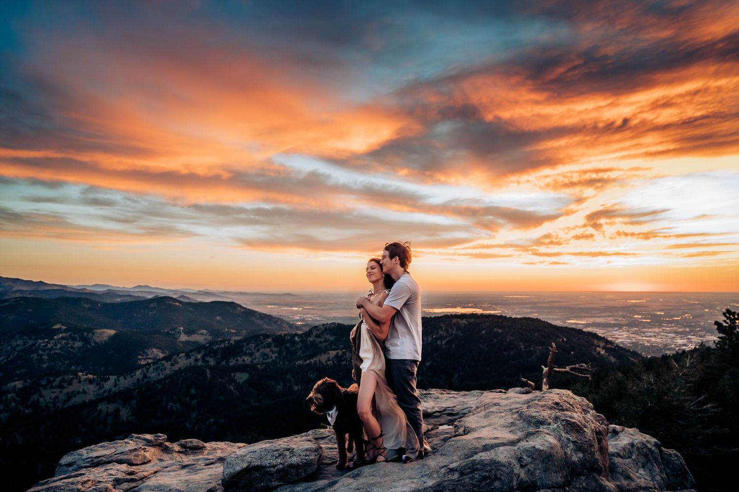 Two silhouettes embrace on a mountain peak against a dramatic orange and purple sunset sky with mountains in background.