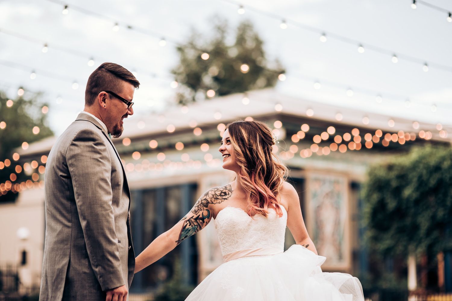 A playful wedding moment captured outdoors with string lights and greenery in the background.