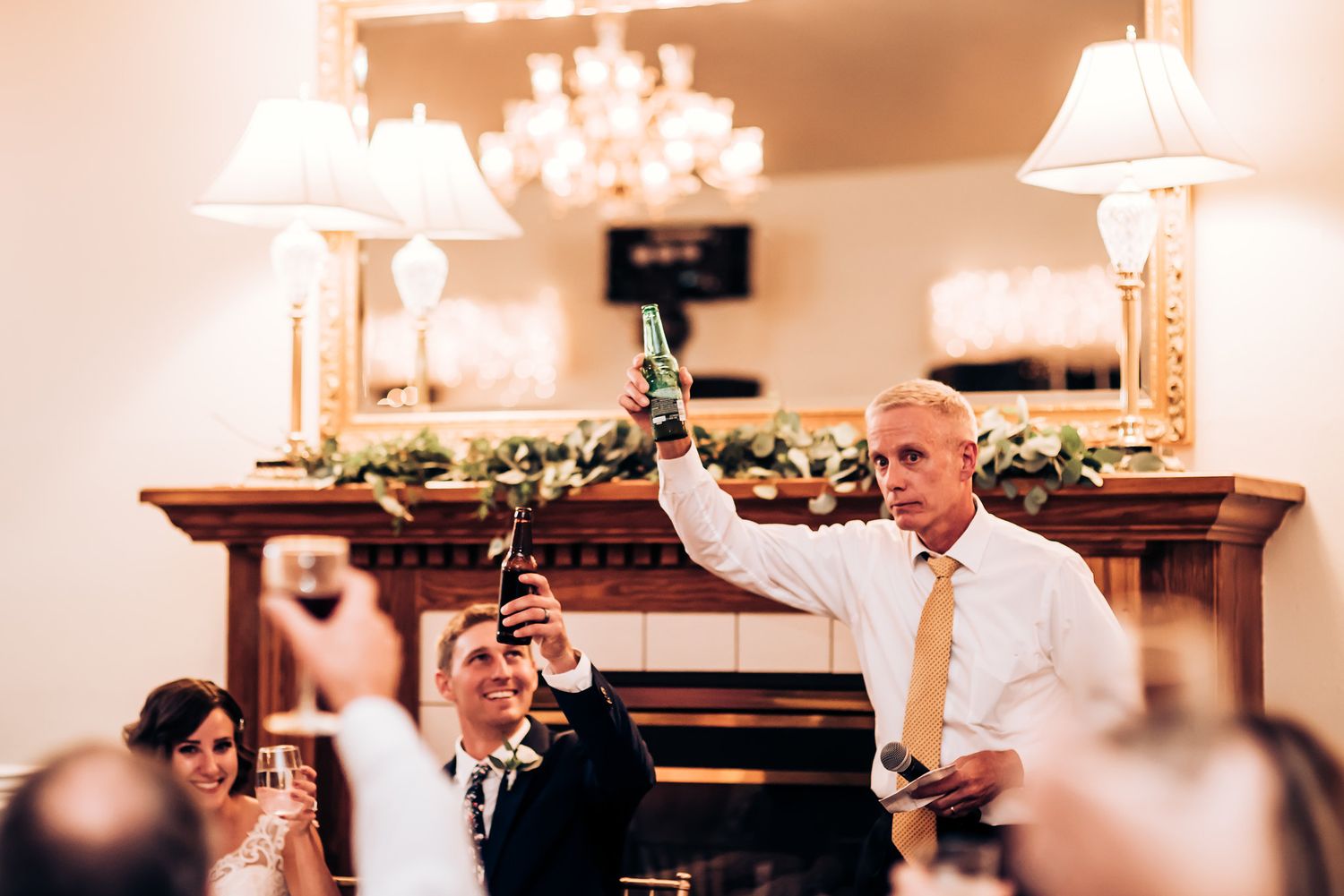 Guest raises champagne glass during celebratory wedding toast near decorated mantelpiece.