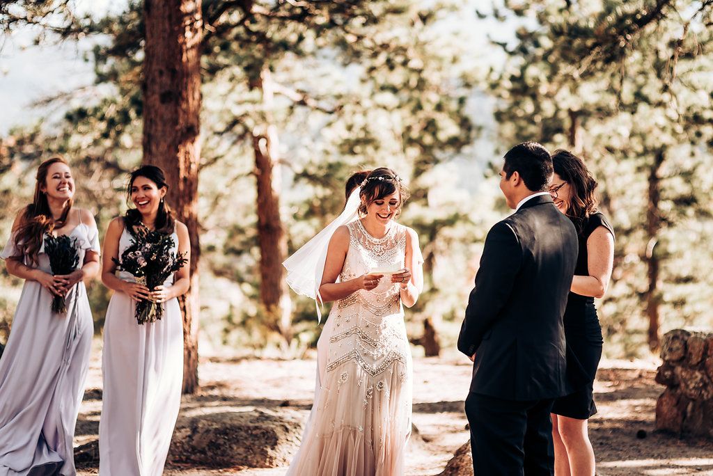A wedding ceremony takes place in a sun-dappled forest with attendants in gray dresses watching the exchange of vows.