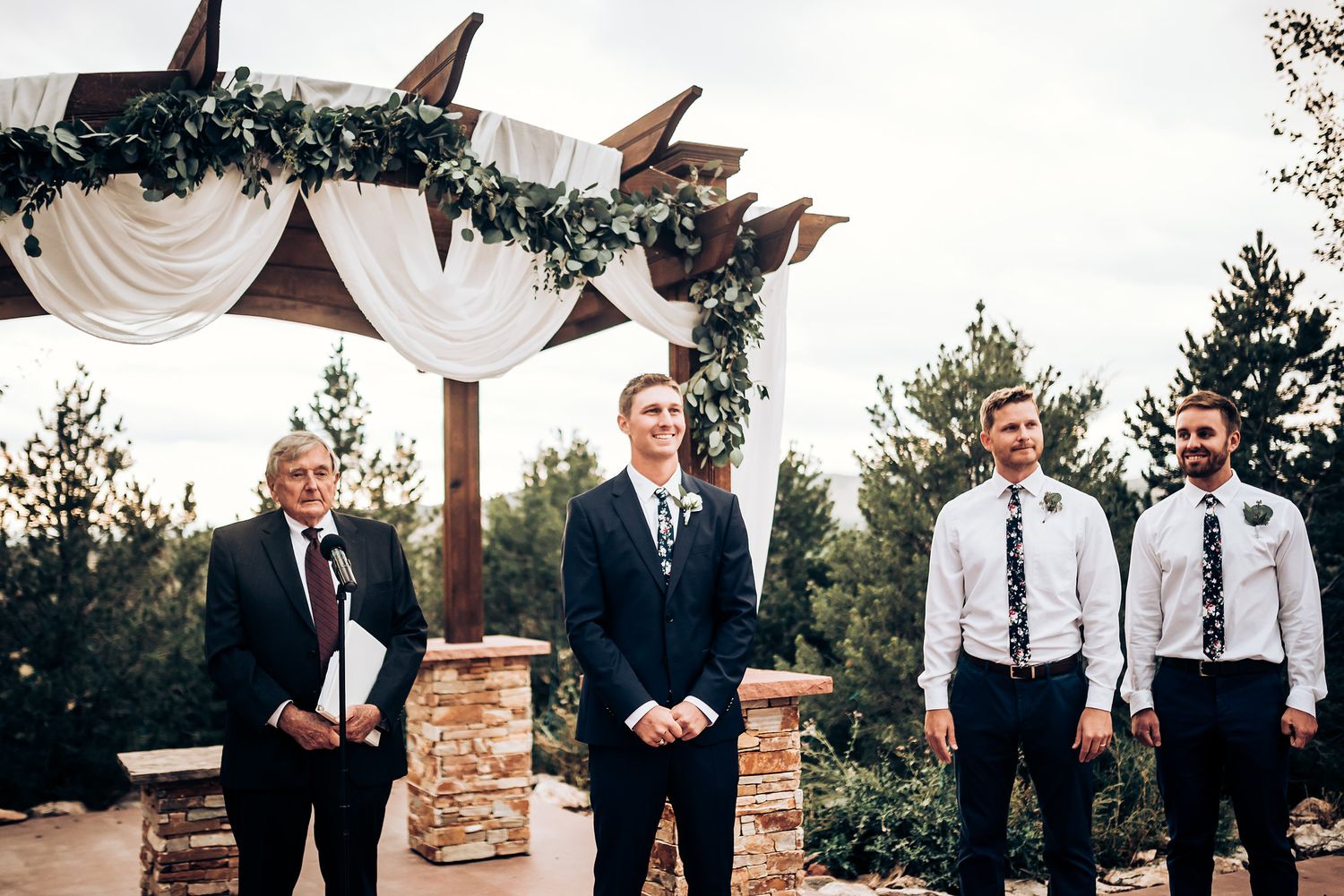 Wedding party members in formal attire stand under a decorated arch with white fabric and greenery.