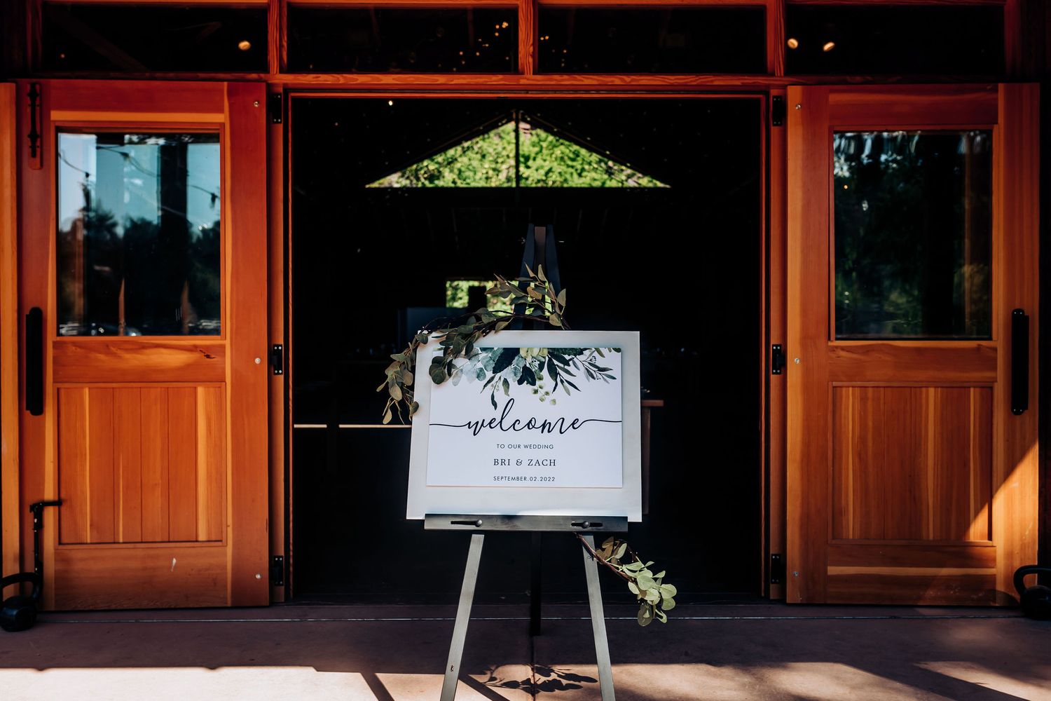 Wedding welcome sign display with greenery at wooden venue entrance.