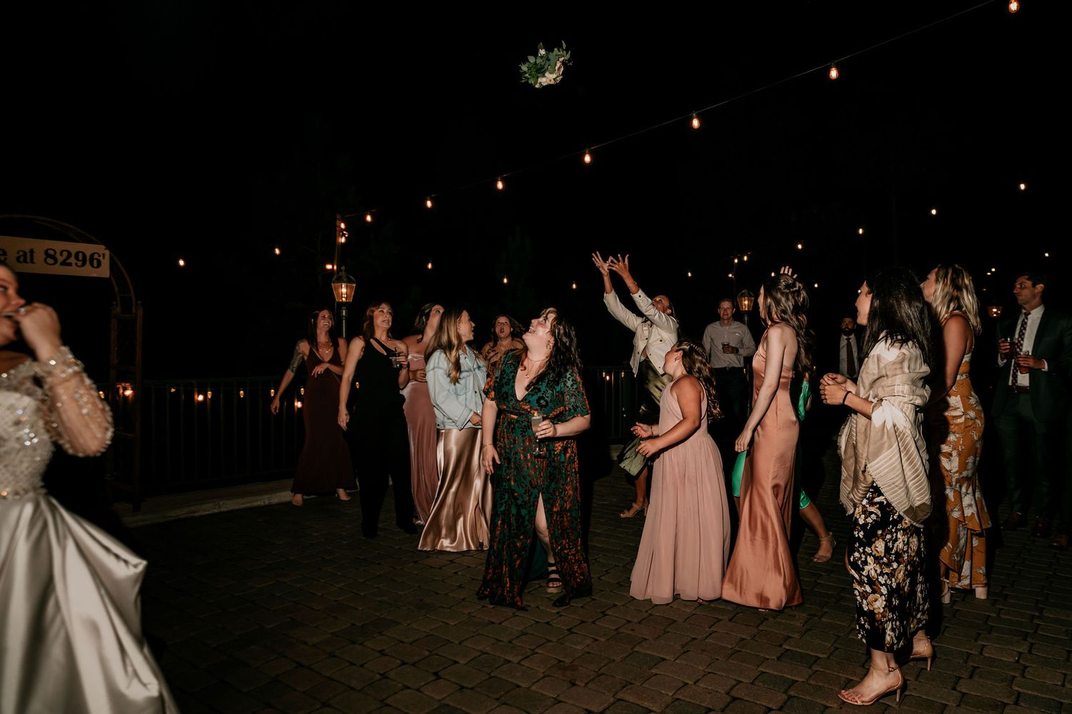 Wedding guests reach for bouquet under the night sky during outdoor celebration.