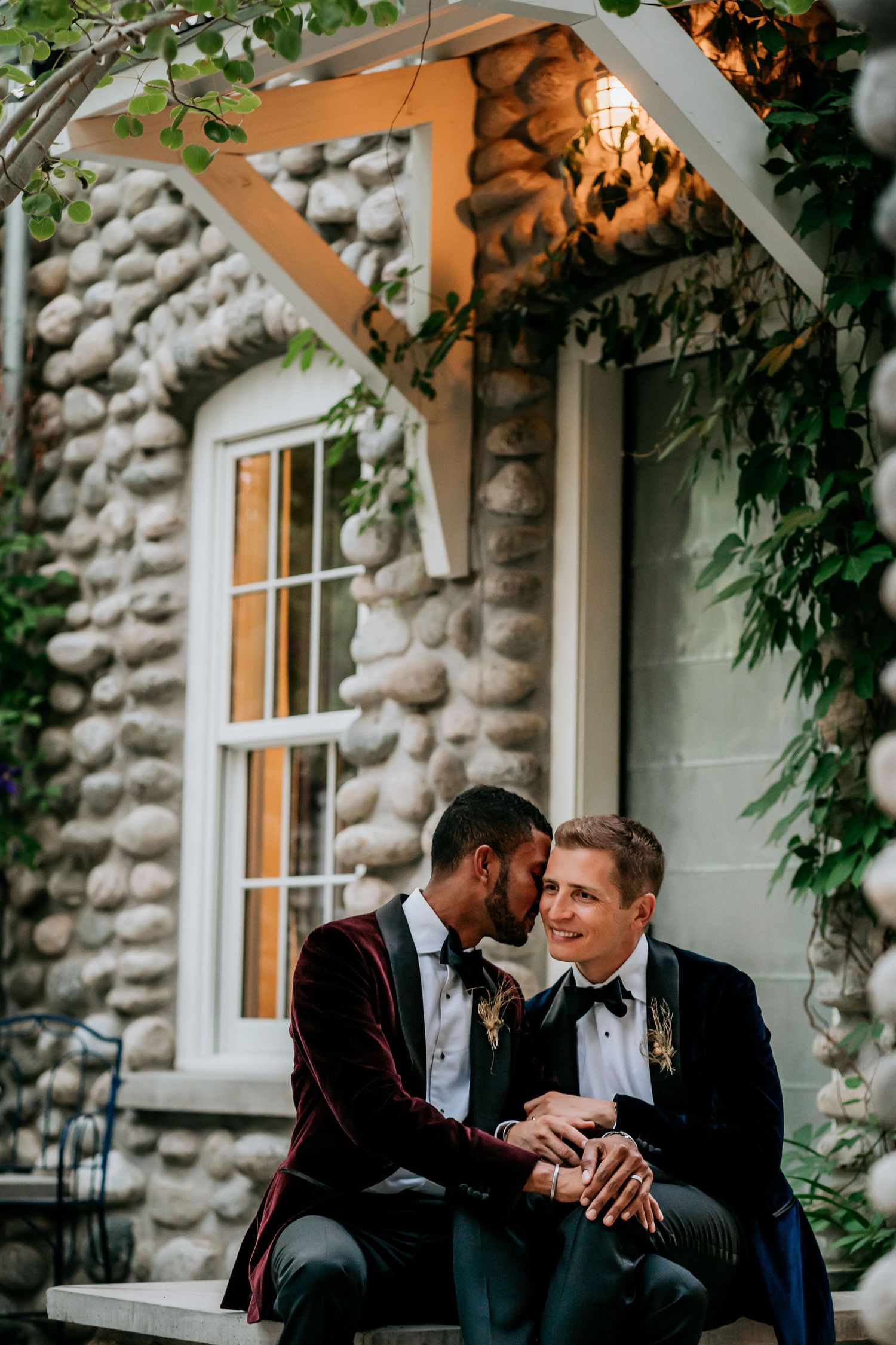 Two people in formal attire share an intimate moment on stone steps of a rustic building.